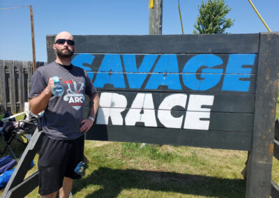 Photo of Sean McCormick showing his medal for completing the Savage Race in front of the Savage Race sign